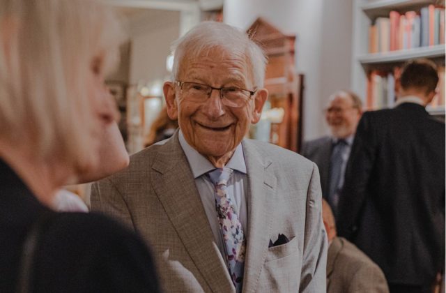Smartly dressed, smiling older man, chatting with a woman.