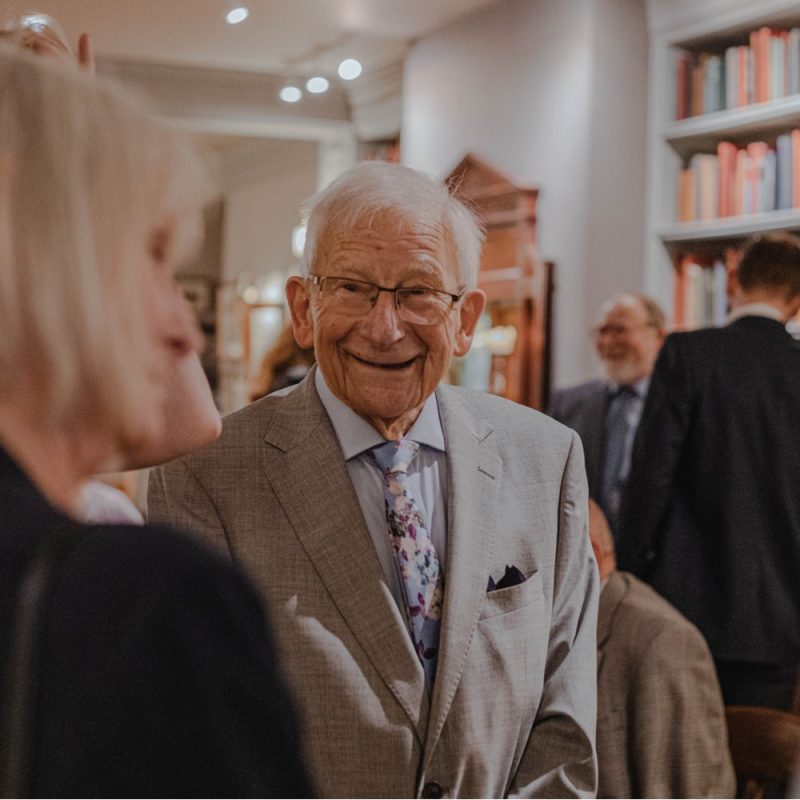 Smartly dressed, smiling older man, chatting with a woman.