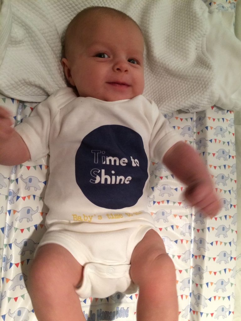 Relaxed looking baby, wearing a 'Time to Shine' branded baby grow.