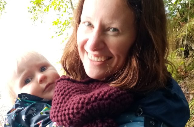 Woman smiling, standing near trees, holding a baby in a sling