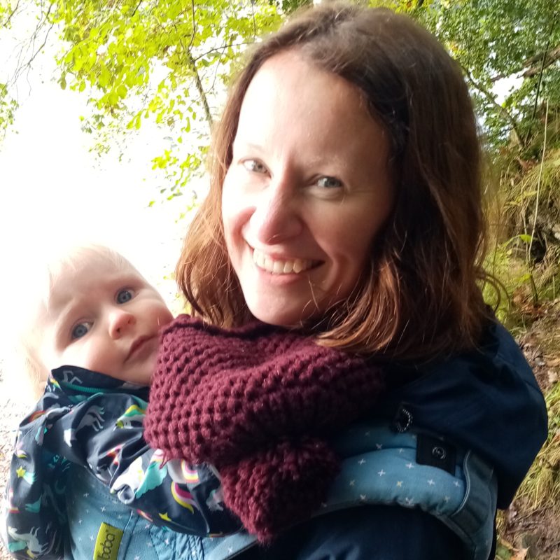 Woman smiling, standing near trees, holding a baby in a sling