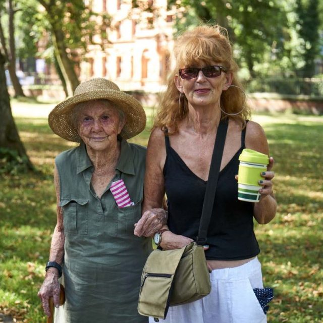 Two women, one much older than the other, walking arm in arm in sunshine.