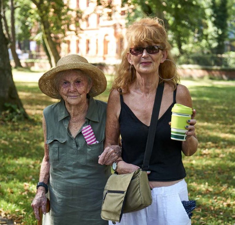 Two women, one much older than the other, walking arm in arm in sunshine.