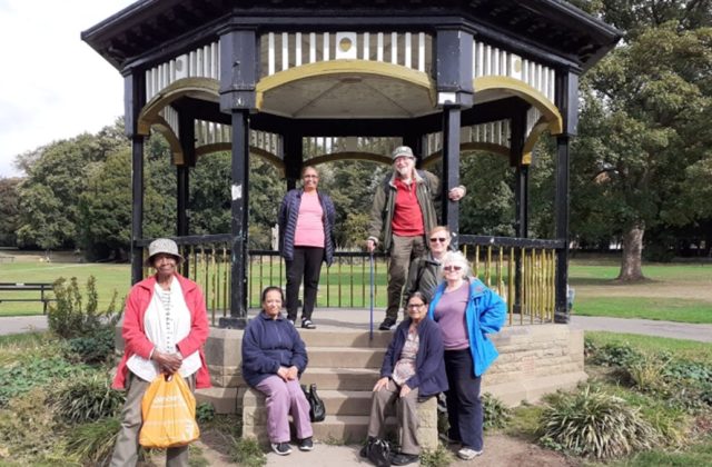Group of 7 older people sat or stood around a park bandstand.
