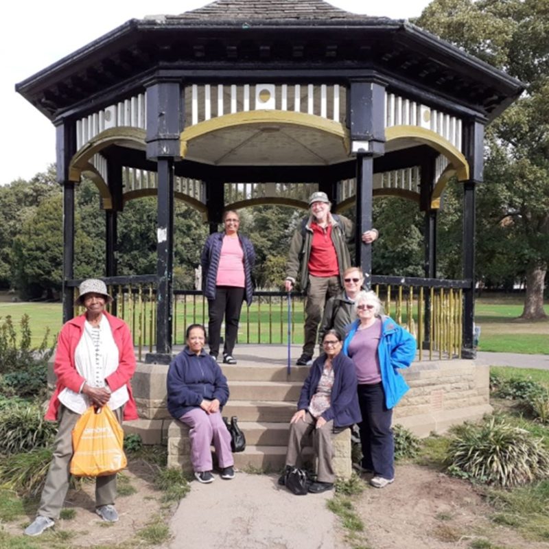 Group of 7 older people sat or stood around a park bandstand.
