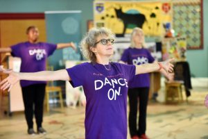 3 women, in a large space, with arms outstretched, wearing purple teeshirts with 'DANCE ON' on front.