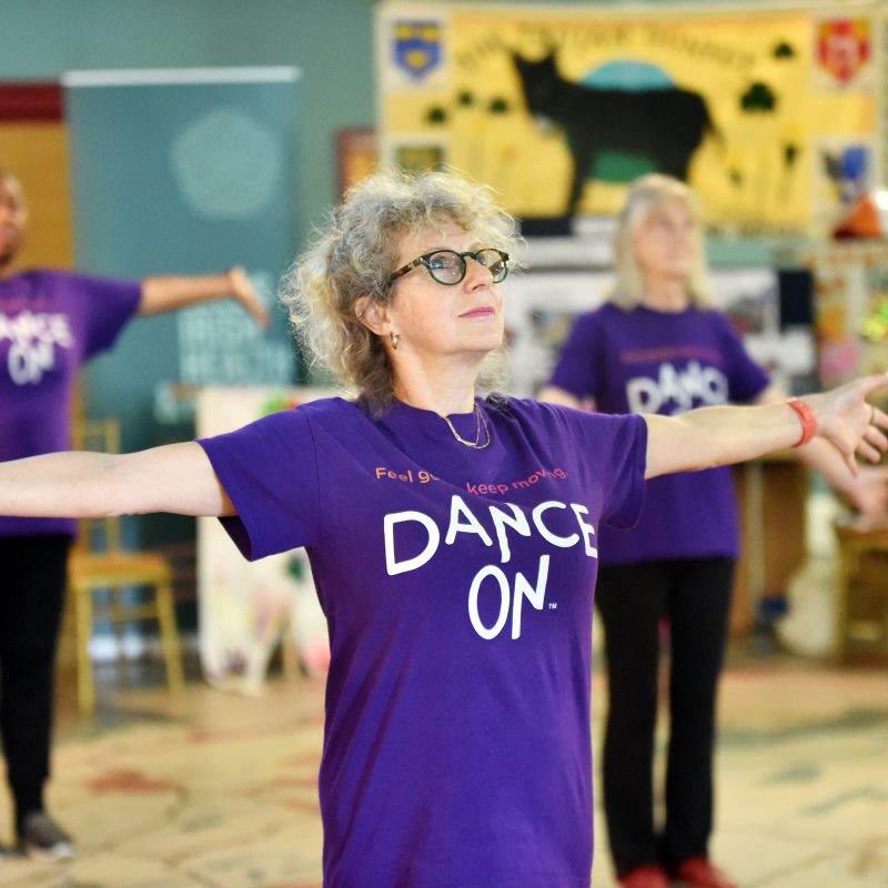 Older women dancers, witharms outstretched, wearing purple teeshirts with 'DANCE ON' on front.