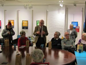 Older man, who is stood up, speaking to a group of mostly older people, sat round a table in a large room.