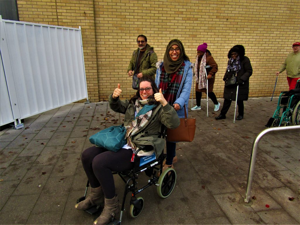 Woman in wheelchair, smiling with both thumbs up, with other people further back, some using walking sticks.