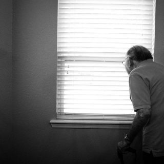Black and white photo. Older man, with walking stick, looking out through blinds