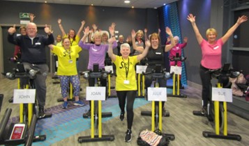 Group of older people, most on static bikes, looking cheerful, each with both arms in the air.