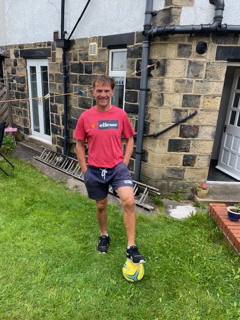 Man standing in a garden, wearing shorts and teeshirt, with his left foot on a football.