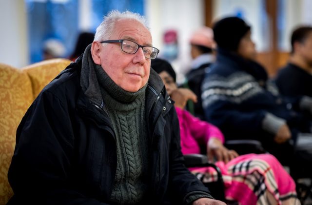Older man, sat inside with coat on, looking at the camera.
