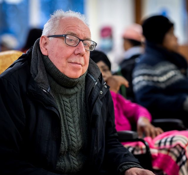 Older man, sat inside with coat on, looking at the camera.