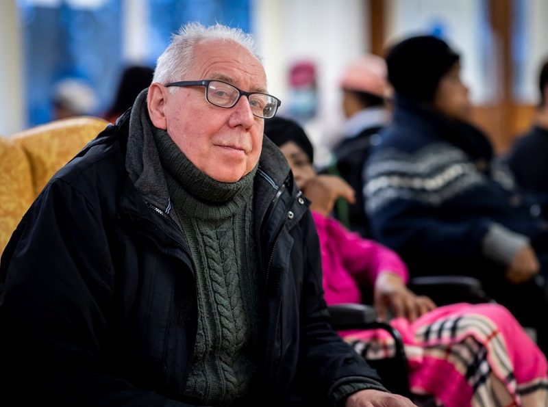 Older man, sat inside with coat on, looking at the camera.