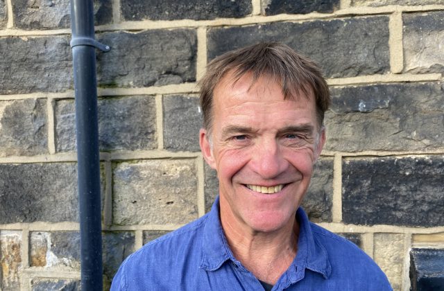 Tony Mullin, wearing a blue shirt, standing in front of a wall