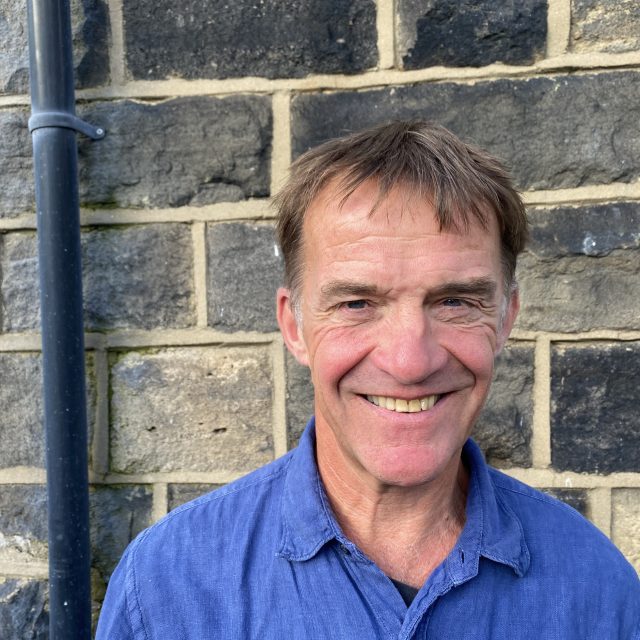 Tony Mullin, wearing a blue shirt, standing in front of a wall