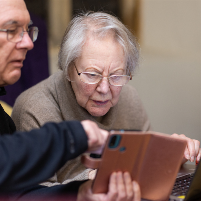 An older man and an older woman looking together at a mobile phone and a laptop.