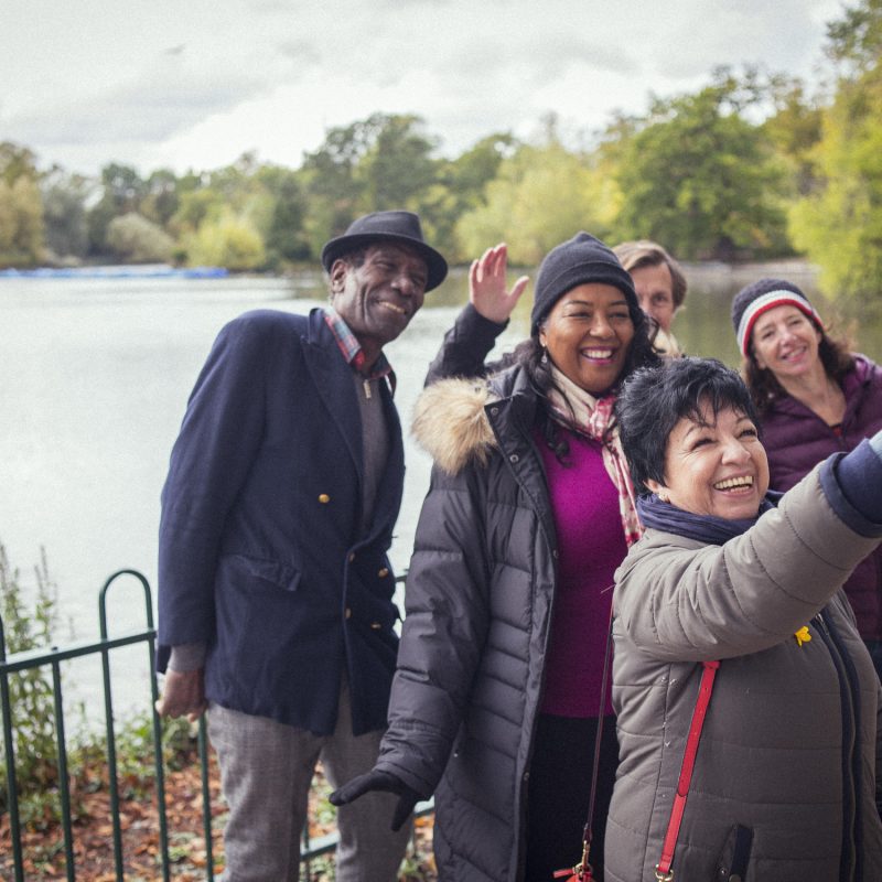 Woman holding up a phone to take a selfie of her, and a group of two other women and two men, who stand behind her. All are smiling and making sure they get into the hot. All are on aa footpath, with a lake behind them..