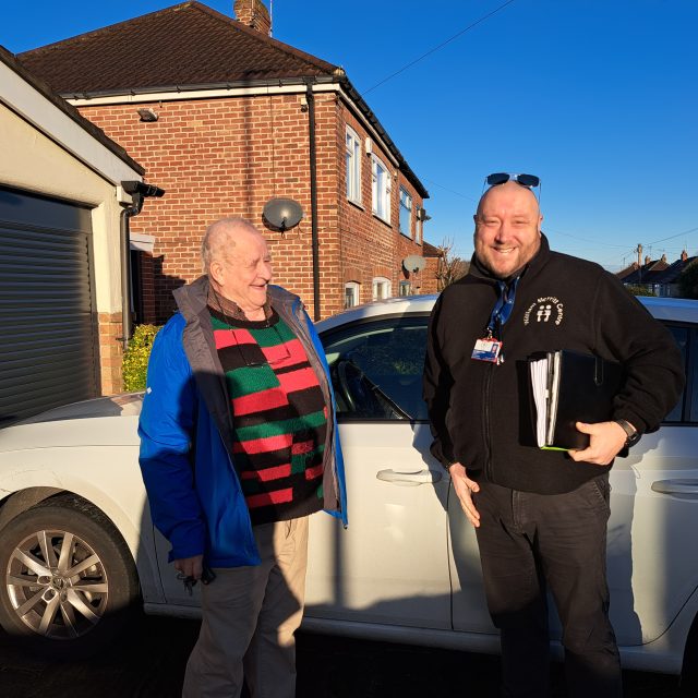 Older man standing with younger man next to a white car.