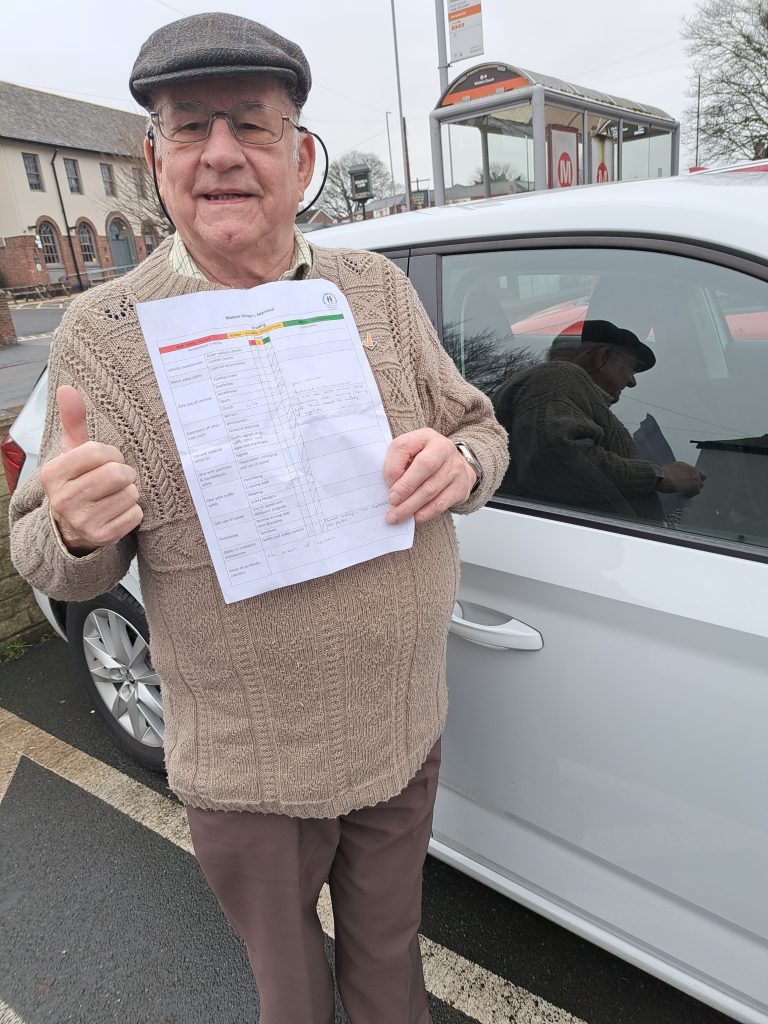Older man standing next to white car, with a thumb's up.He is holding a piece of paper in his left hand.