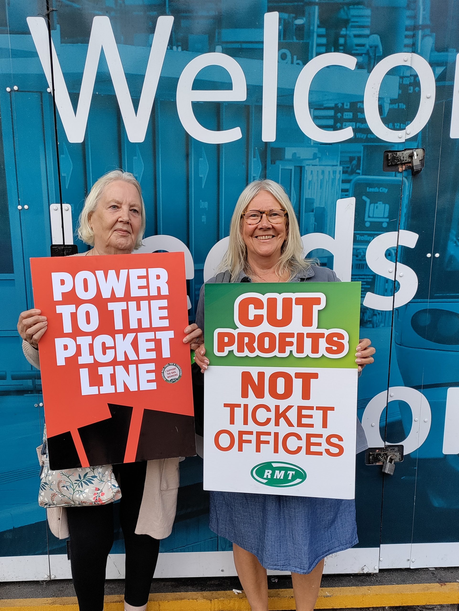 Two women stood next to each other, each holding a placard in front of them. Text on the placards: "Power to the picket line" and "Cut profits not ticket offices - RMT"