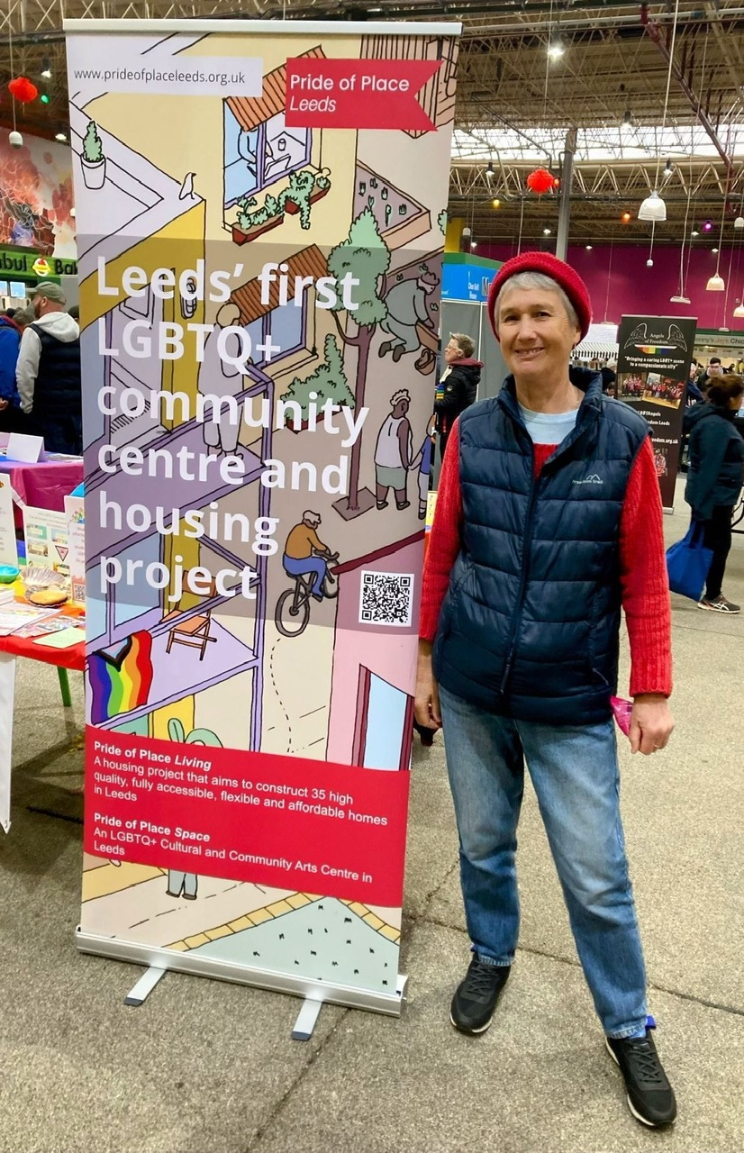Woman standing, smiling, next to a colourful pop-up banner with main text "Leeds' first LGBTQ+ communitty centre and housing project"
