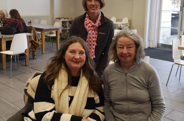 Three women smiling at camera, two sat in front of the other, who is standing.