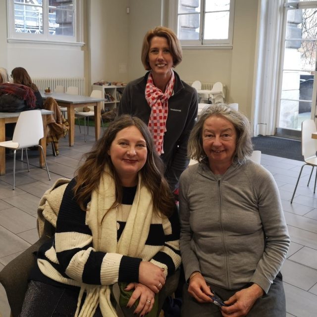 Three women smiling at camera, two sat in front of the other, who is standing.