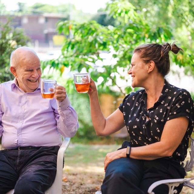 An older man and a younger woman sat with drinks in a cheers motion